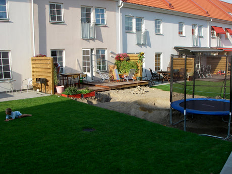 Radhusträdgård under anläggning med grushög och gropar för damm, träd och studsmatta – compact gardening på 6,5×10 m med planerat trädäck, gräsmatta, blommor, kryddväxter och bärbuskar i sol och skugga.
