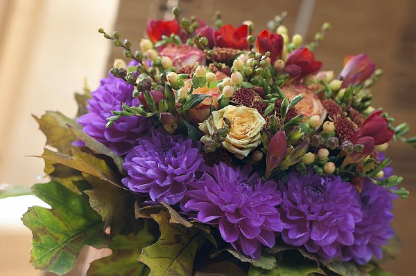 Close-up of a Scandinavian Forest Craft bouquet: cut flowers (rose, dahlia, chrysanthemum, lily, freesia) in mossy green and earthy brown with pink, red, and yellow accents; handcrafted, Swedish-heritage design inspired by Från Blomsterfrämjandet.