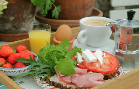 Frukost i det gröna i juni i Tyras växthus och trädgård: ljus morgon, potager med rucola och våtarv, tomatplantor som tjuvas, doft av rosor, syren och kaprifol.